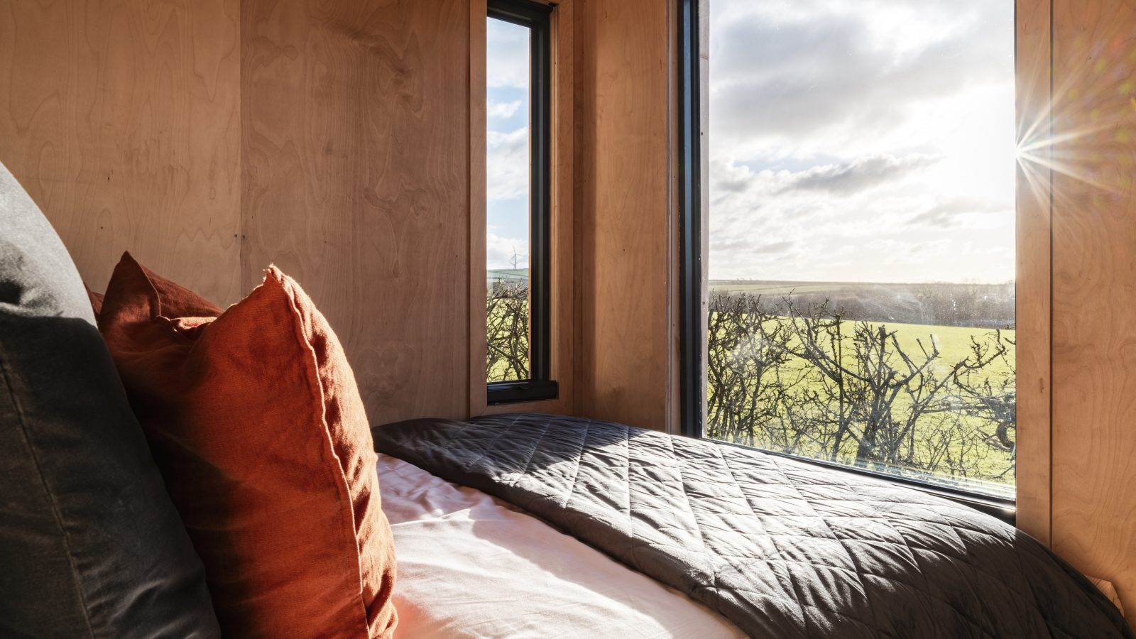 A cozy bed with black and rust-colored pillows is positioned next to two large windows at The Lookout, one of which has sunlight streaming through. The windows offer a scenic view of Bogee Farm's green field and distant trees under a partially cloudy sky.