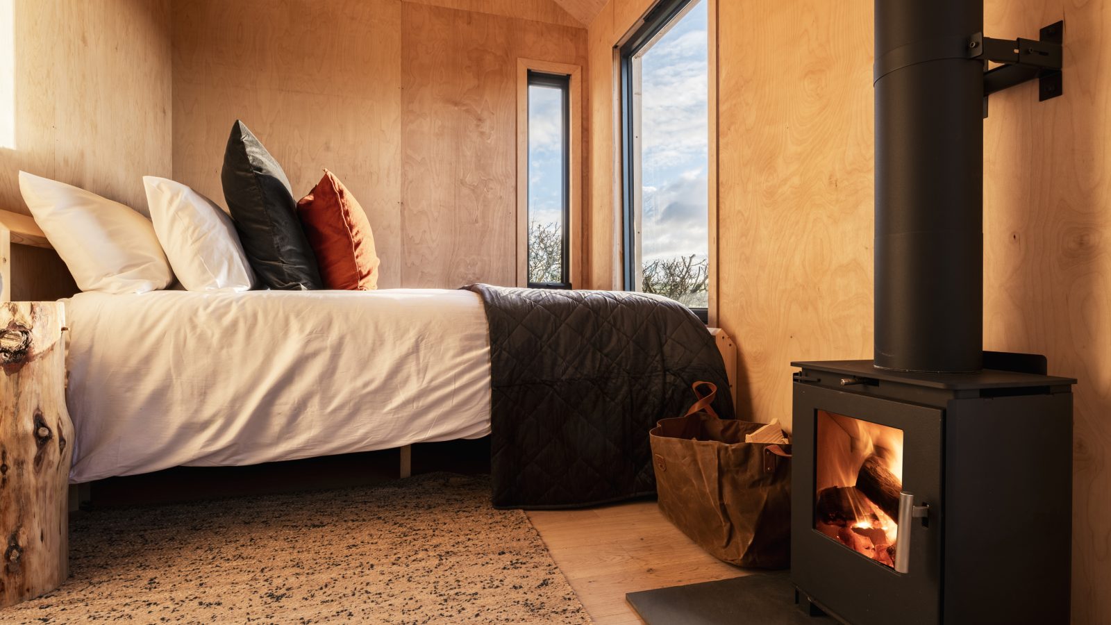 A cozy wooden cabin interior at The Lookout features a neatly made bed with pillows and a dark quilt. A wood-burning stove with a small fire burning inside is on the right. The large windows allow natural light to fill the space, highlighting the warm wooden tones.