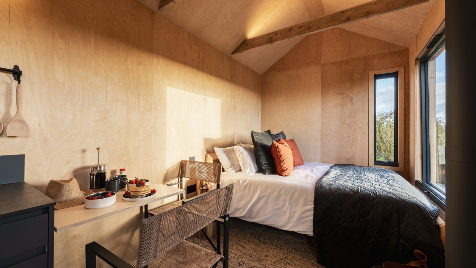 A cozy, wood-paneled tiny house interior at Bogee Farm features a bed on the right, adorned with pillows and a black comforter. A small table on the left displays breakfast items like toast, coffee, and berries. A woven chair sits at The Lookout's table, and a tall window lets in natural light.