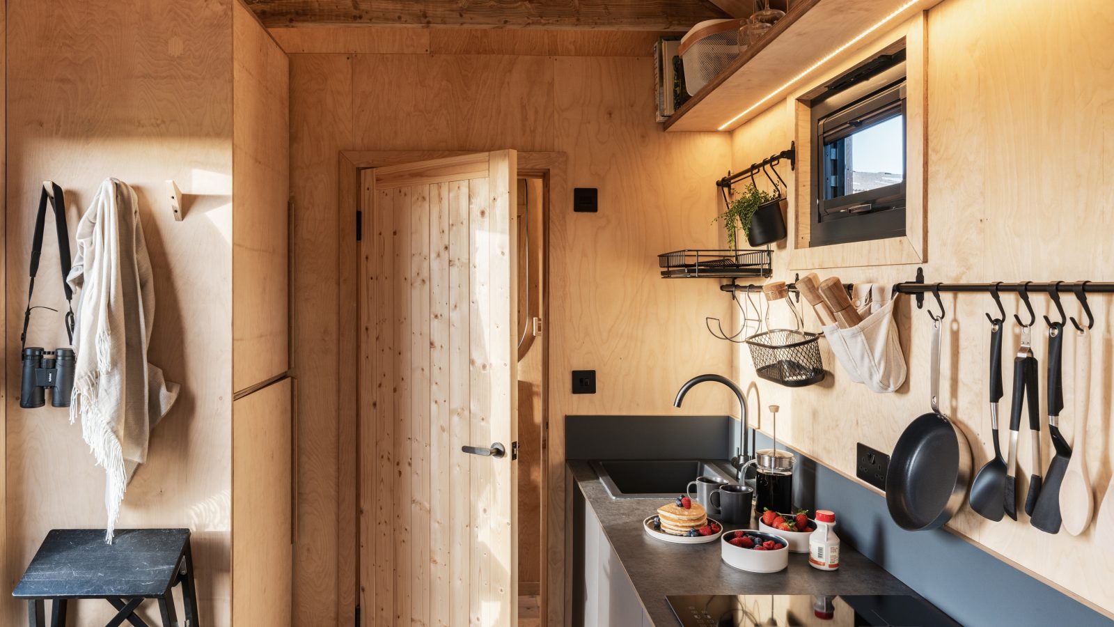 A compact wooden kitchen at Bogee Farm with modern amenities. It features a sleek stovetop, cooking utensils hung on a rail, hanging plants, shelves with dishes, and a small window. A door in the center leads to another space, while a stool and hanging fabric are on the left.