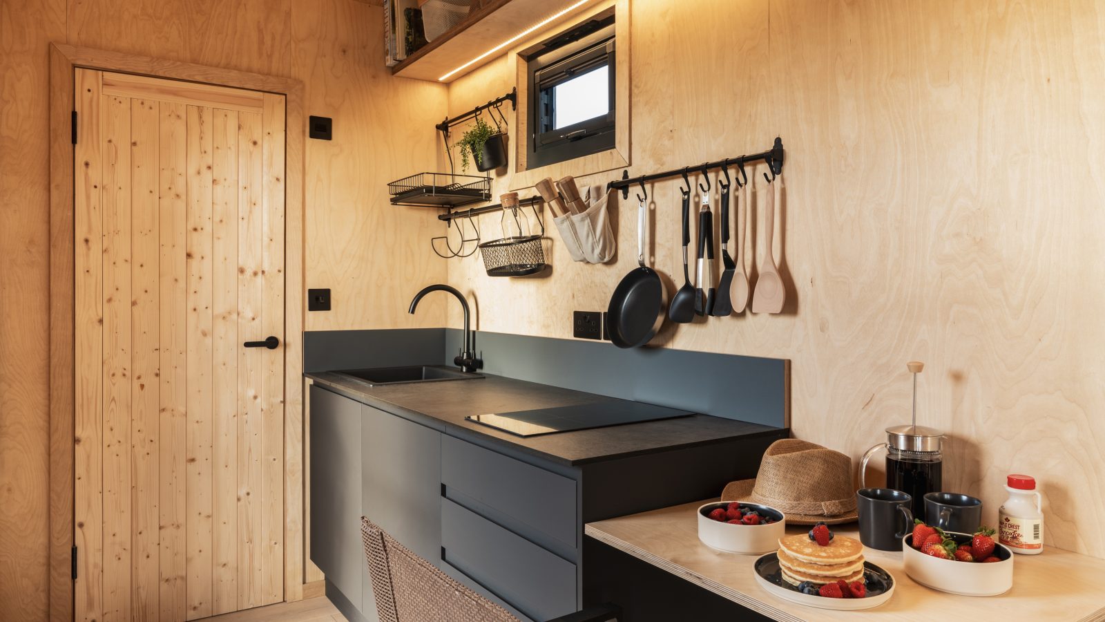 A cozy kitchen at Bogee Farm features wooden walls and a wooden door on the left. The black countertop has a sink and cooking utensils hanging above it. On a small table, there's a plate of pancakes with berries, syrup, and various fruits—a perfect spot for a rustic lookout.