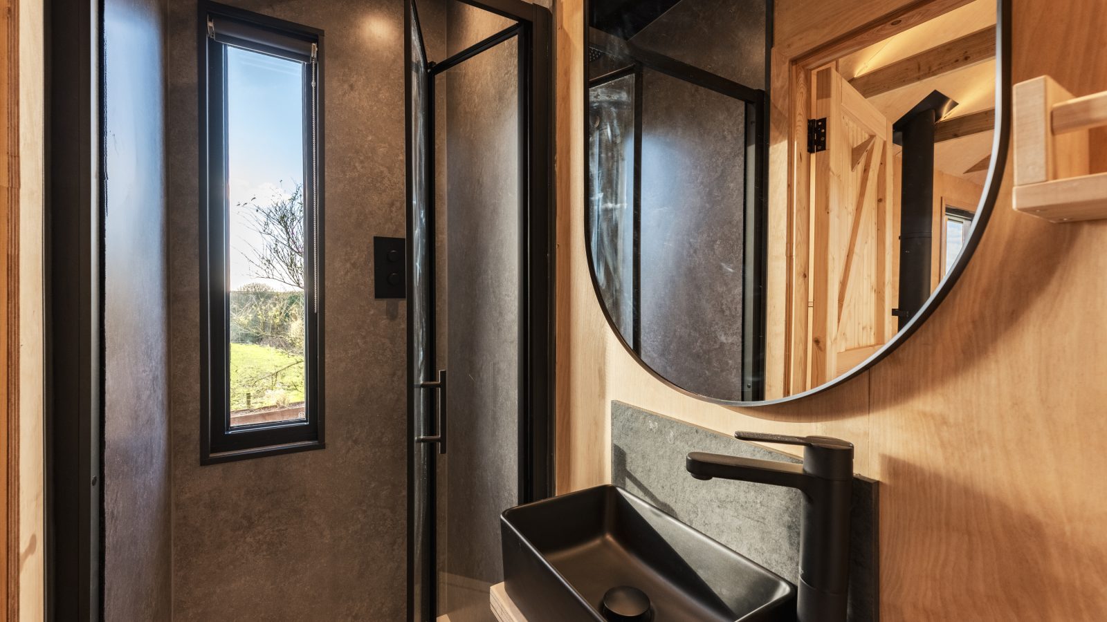 Modern bathroom with a round mirror, black basin, and sleek black faucet. The shower area has glass doors and a tall rectangular window letting in natural light, offering a lookout over Bogee Farm's lush greenery. Light wood and black accents create a contemporary look.