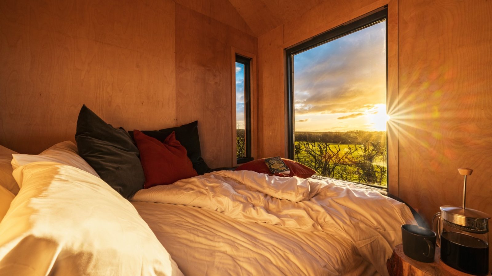 A cozy wooden bedroom at Bogee Farm with a large bed adorned with white and black pillows and a white comforter. The room features two large windows overlooking the scenic landscape of The Lookout during a radiant sunset. A small wooden table holding a French press and two cups is beside the bed.