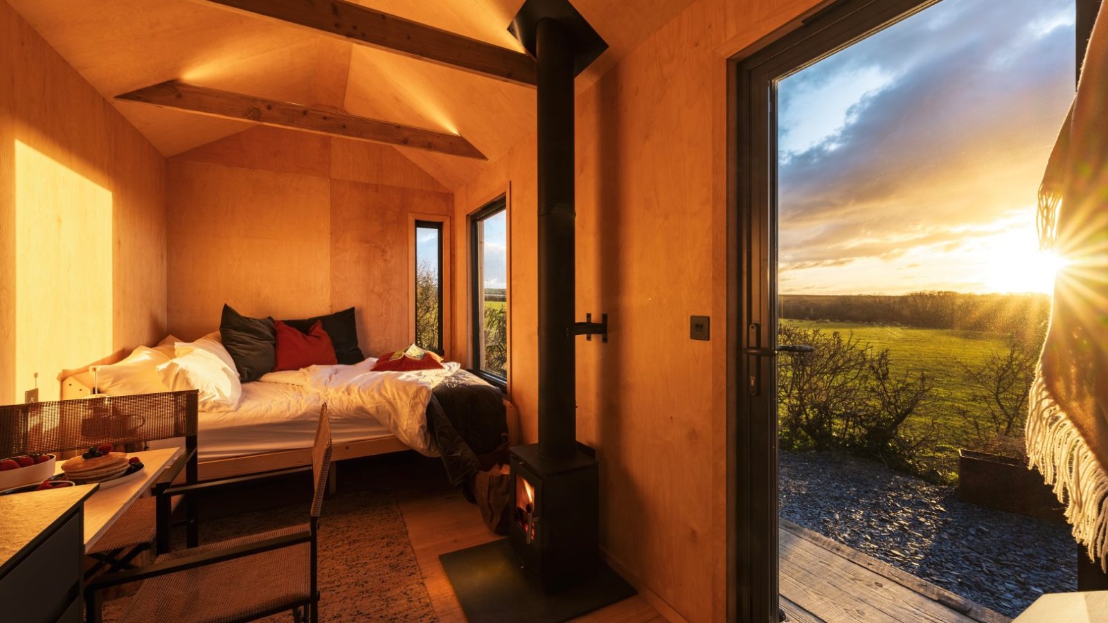 A cozy wooden cabin interior at The Lookout features warm lighting, a neatly made bed, a wood-burning stove, and a small table with a chair. Large windows and a glass door reveal a stunning sunset over Bogee Farm's green landscape. A blanket is draped over a chair near the doorway.