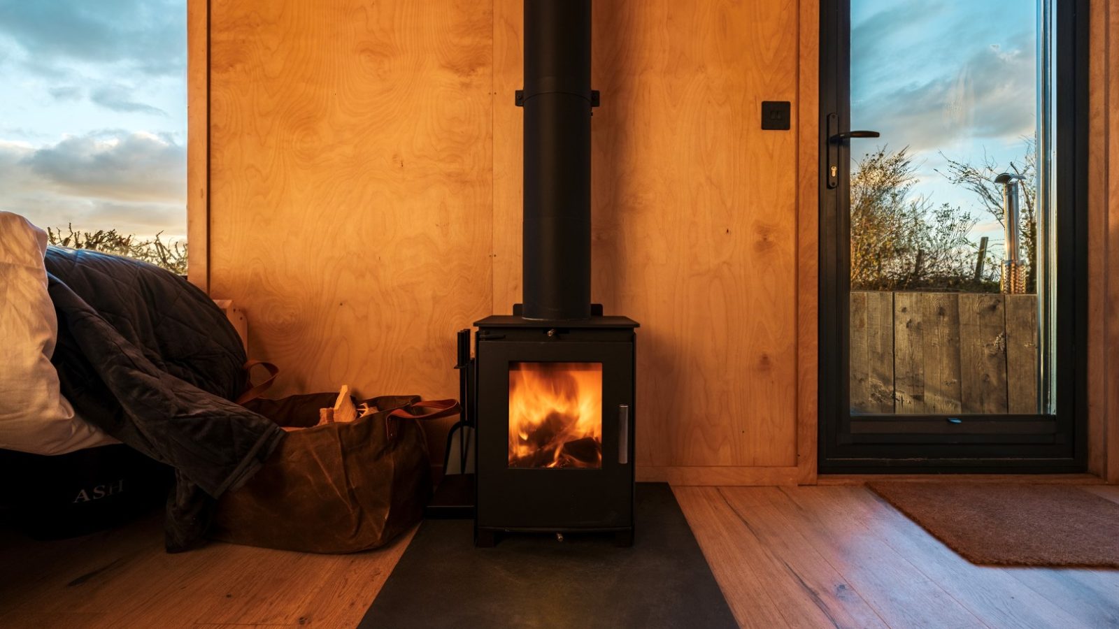 A small black wood stove with a fire burning inside is situated against a wooden wall in a cozy room at Bogee Farm. To the left is a bed with a bag beside it. A door on the right opens to an outdoor view of a sunset sky, giving the room, which serves as an ideal lookout, a warm, inviting atmosphere.