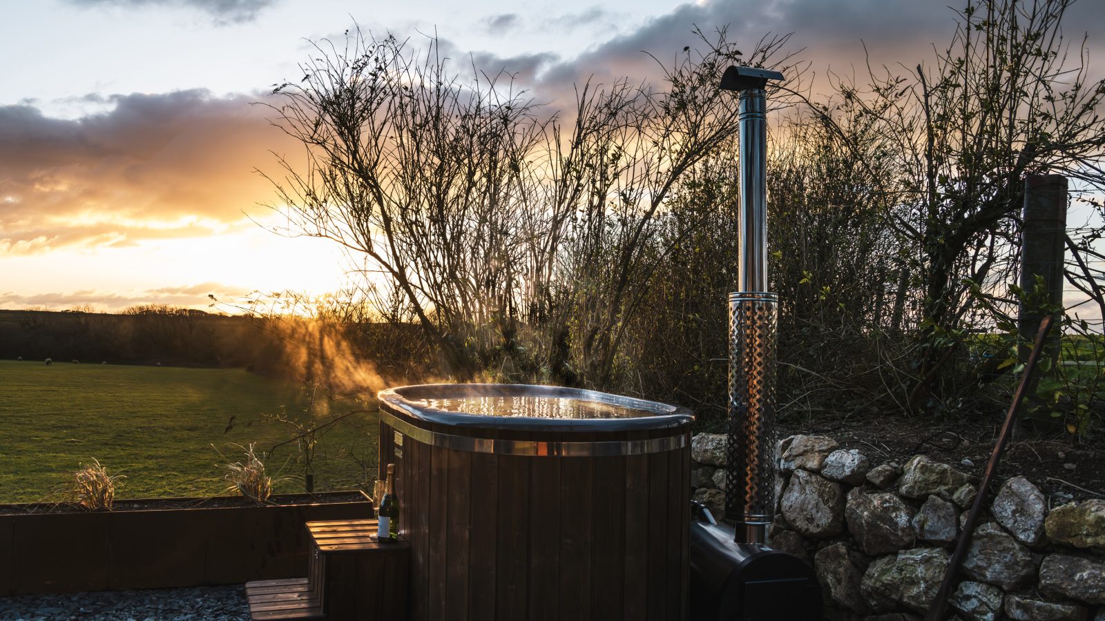 A wooden hot tub with steam rising sits outdoors near a stone wall at Bogee Farm, against a backdrop of a grassy field and leafless trees at sunset. The sky has a warm orange hue, and a small table with a drink is placed beside the tub for the perfect lookout experience.