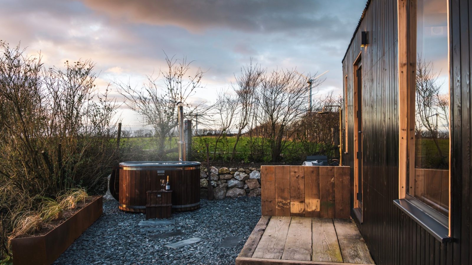 Outdoor setting at The Lookout with a wooden hot tub beside a small, dark-colored building. There is a wooden bench and planter boxes filled with tall grasses. The scene at Bogee Farm is set against a backdrop of bare trees and a cloudy sky, suggesting a cool, peaceful atmosphere.