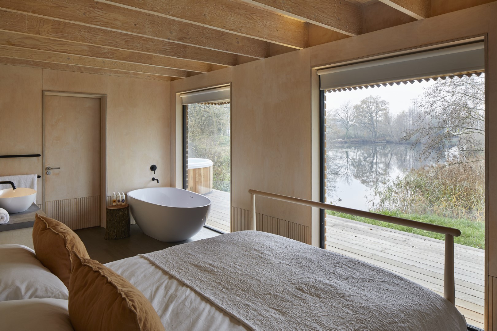 A luxury lakeside cabin just 2 hours of London, features a cosy bed with tan pillows and a freestanding bathtub by large windows overlooking a serene lakeside view. Natural wood walls and ceiling beams frame the space, creating a warm, inviting atmosphere. A deck extends outside, enhancing the outdoor connection.