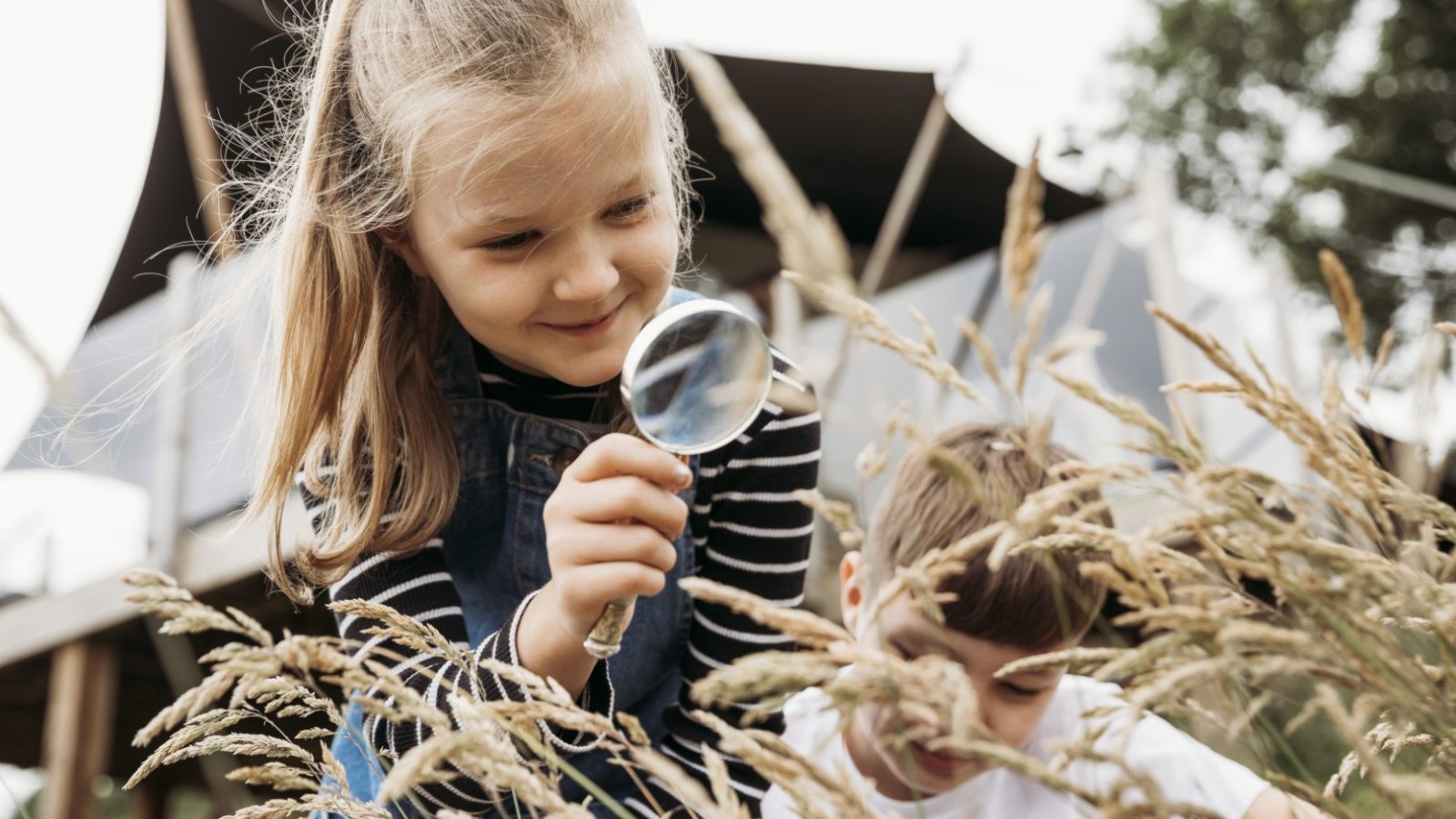 A young girl with a ponytail holds a magnifying glass, examining tall grass intently, while a boy looks down at something beside her. They are outdoors near a safari tent, with a building structure and trees in the background. The scene appears lively and investigative.