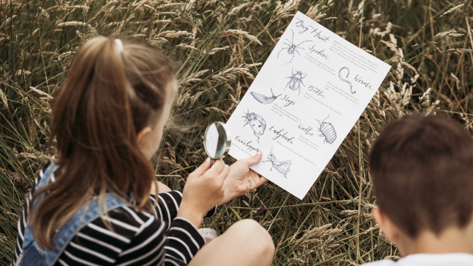 Two children sit in a grassy field, near a safari tent, examining a nature scavenger hunt sheet. One holds a magnifying glass and the sheet with illustrations and names of insects and plants. The other child’s back is partially visible as they look at the sheet together.