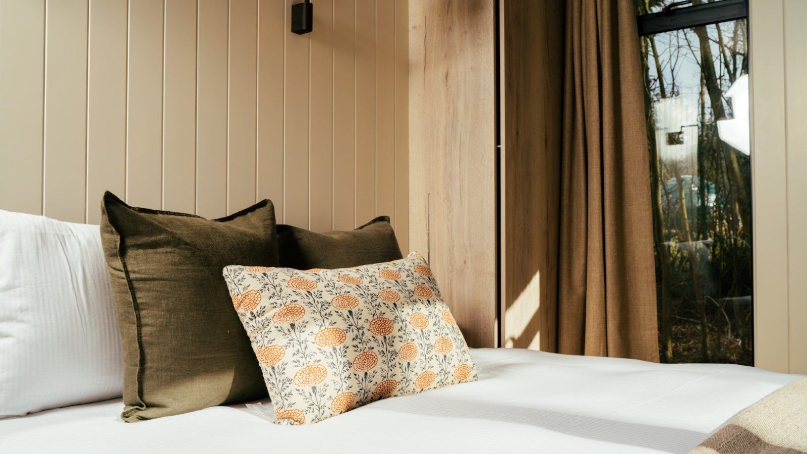 A cozy bedroom at Camp Katur features a bed with white linens, topped with green and floral-patterned pillows. Neutral-toned walls, wood paneling, and a window with beige curtains allow natural light to filter through, creating a calm and inviting atmosphere perfect for glamping enthusiasts.