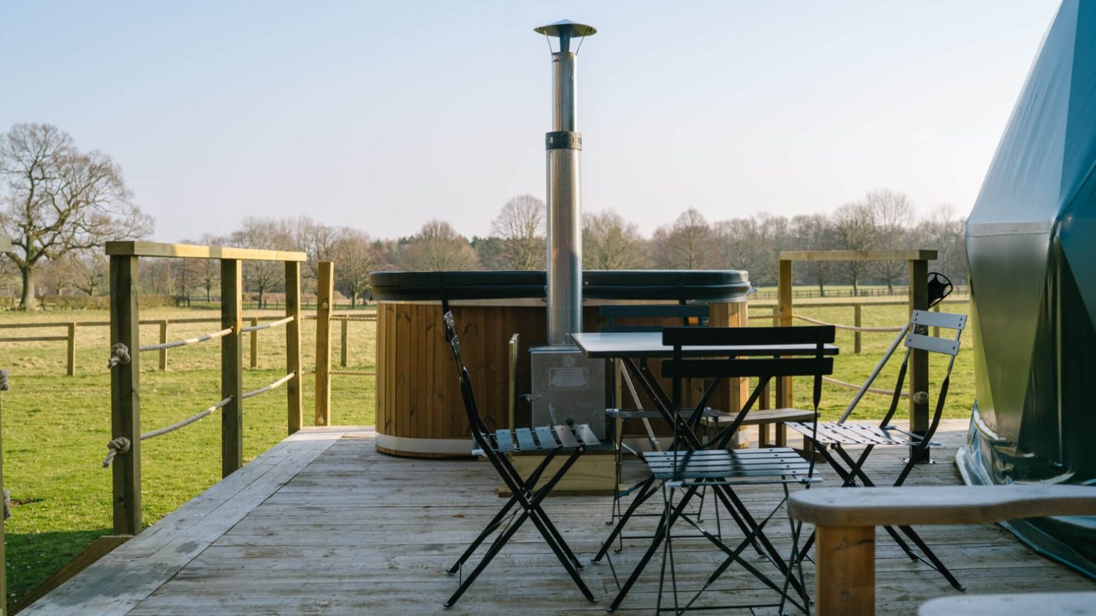 A wooden deck with a hot tub, metal table, and chairs overlooks a grassy field surrounded by a fence. The setup includes a chimney attached to the hot tub and is under a clear blue sky with trees in the background, perfect for Glamping at Camp Katur and enjoying outdoor activities.