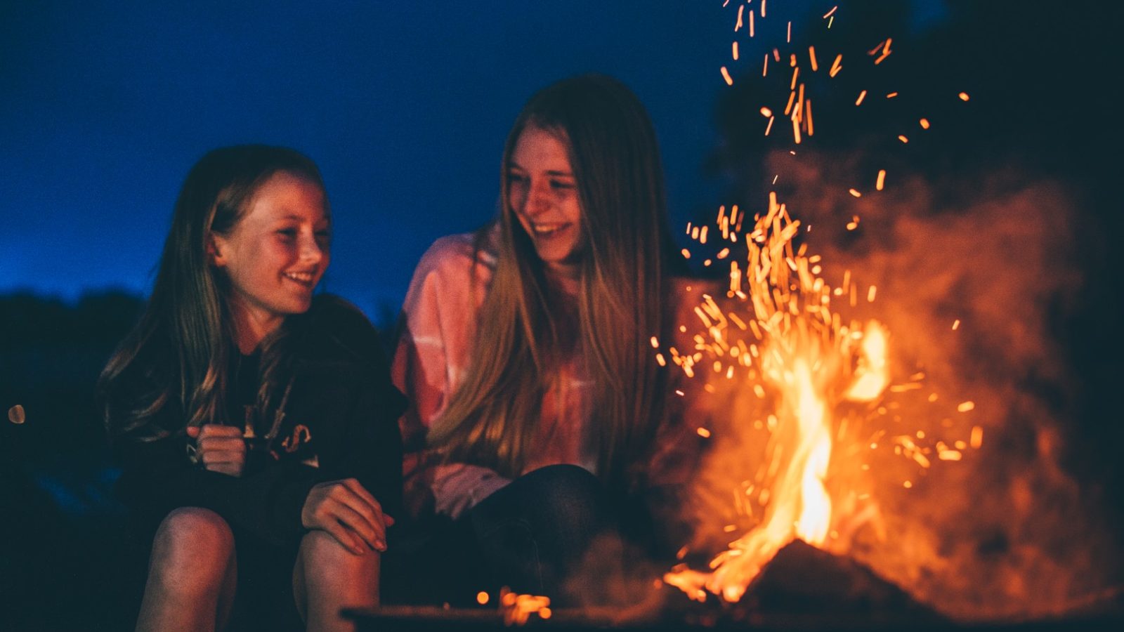 Two girls sit around a campfire at night, smiling and talking. Sparks from the fire light up their faces, creating a warm glow against the dark sky in the background. They appear to be enjoying a peaceful moment outdoors, perhaps sharing stories of Nantseren adventures.