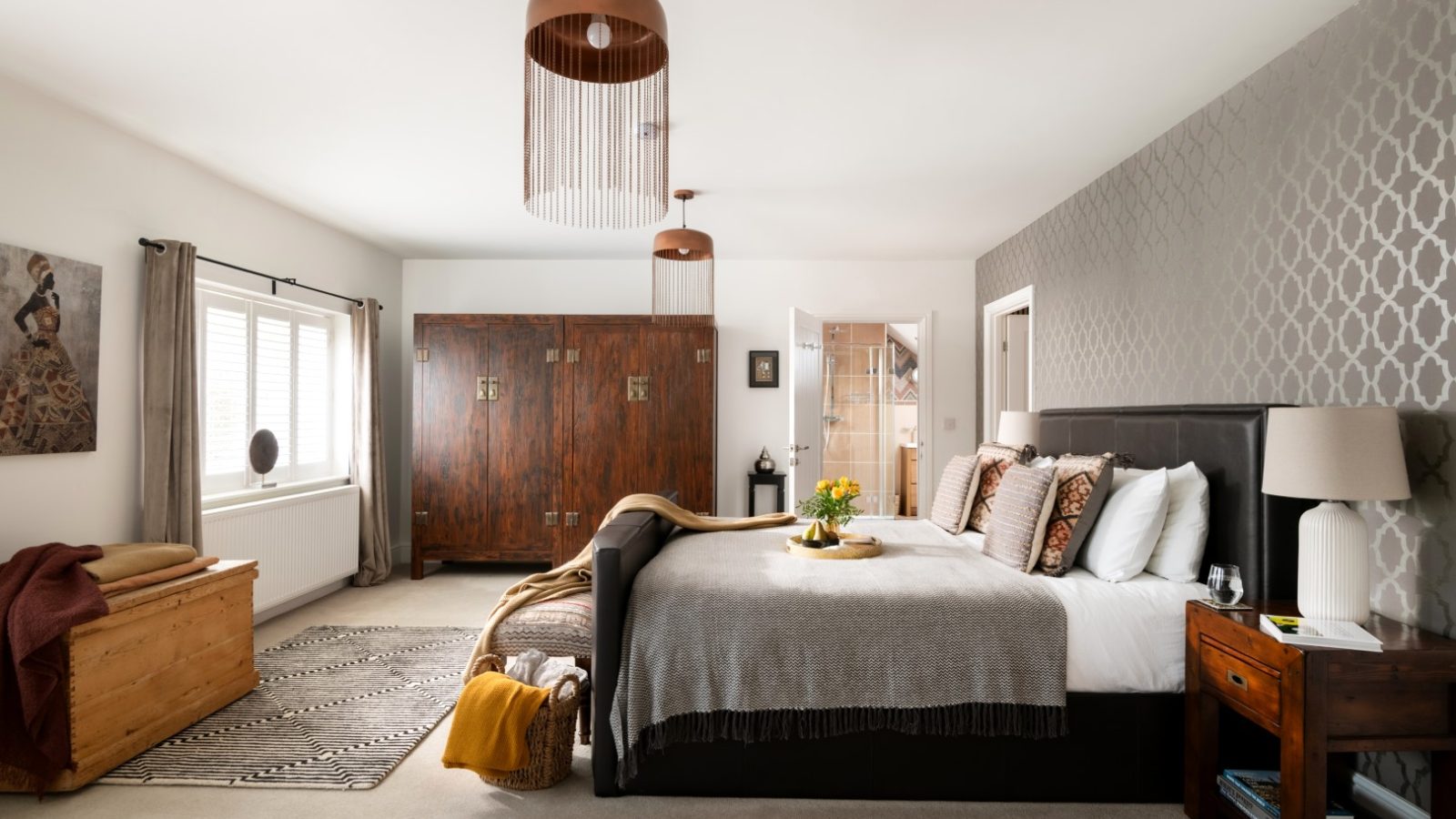 A spacious West Acre bedroom with a large bed adorned with pillows and a gray blanket. The room features wooden furniture, including a wardrobe, chest, and two nightstands. A doorway leads to a bathroom. Pendant lights hang from the ceiling, and a patterned accent wall adds texture.