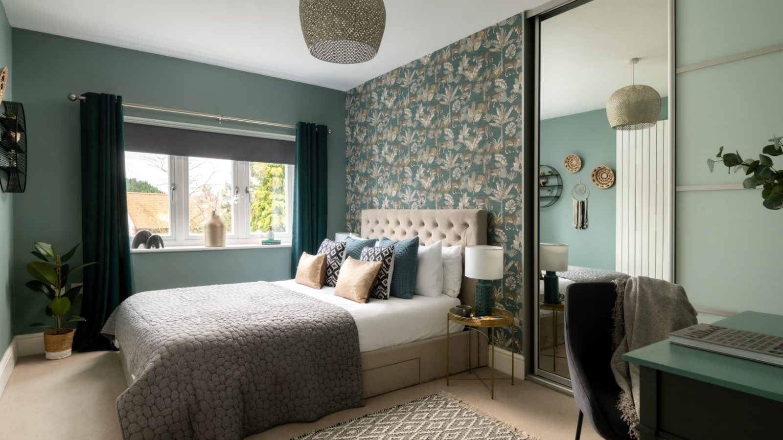 A serene West Acre bedroom with teal walls and a patterned accent wall behind a tufted beige headboard. The bed has gray bedding and assorted pillows. A large mirror-door wardrobe, a window with dark curtains, a desk with a chair, and a potted plant complete the room.