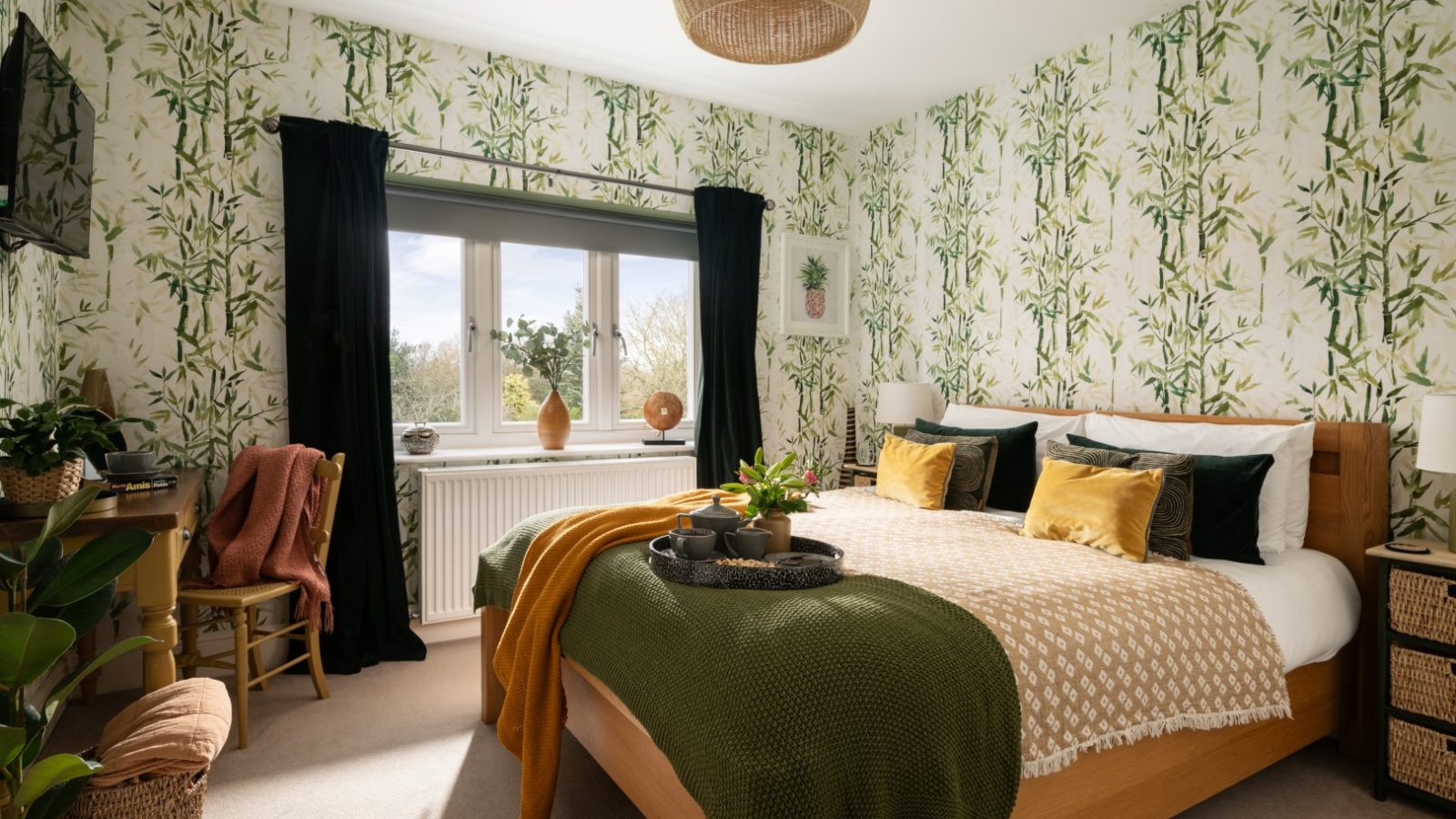 A cozy bedroom in West Acre with a double bed featuring green and yellow pillows, a green throw blanket, and a beige comforter. The walls are decorated with leafy wallpaper, and a large window with black curtains allows natural light in. The room includes plants and wicker decor, enhancing the tranquil vibe.