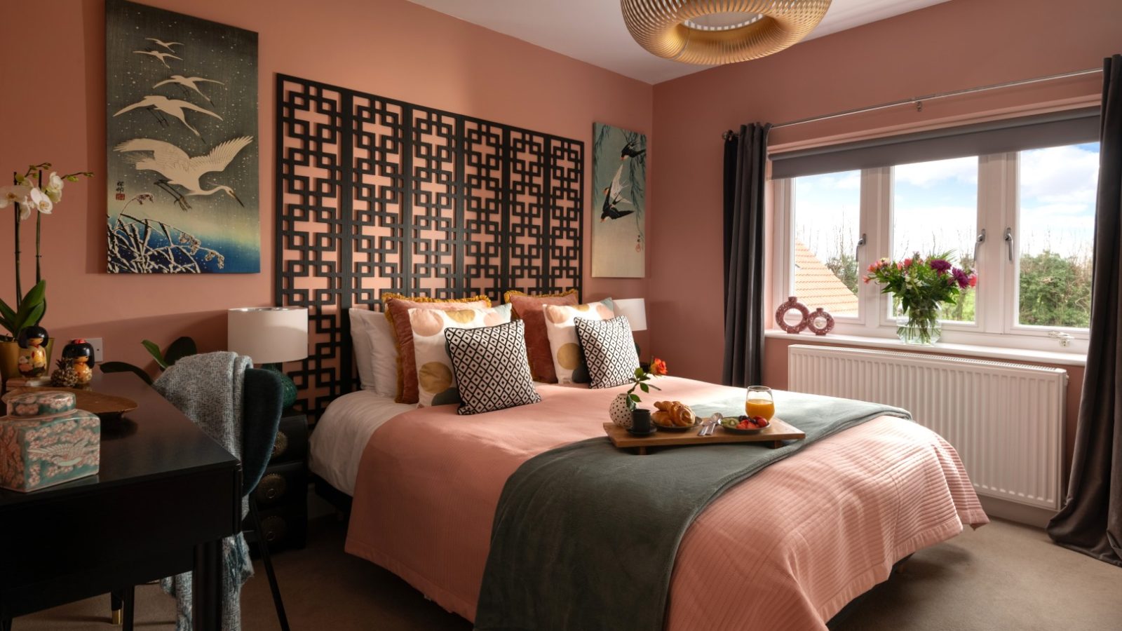 A cozy bedroom in West Acre features a pink accent wall with intricate designs and bird artwork. The bed is neatly made with pink and green bedding, adorned with stylish pillows. A wooden tray with breakfast sits on the bed. Large windows let in natural light, and a modern chandelier hangs above.