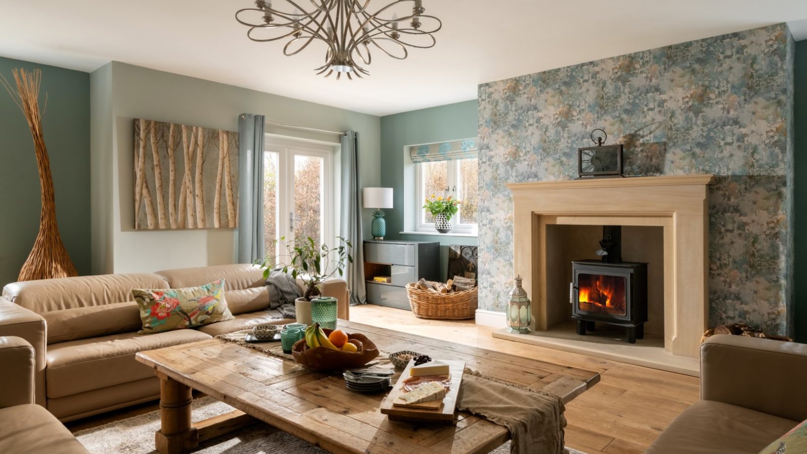 The cozy West Acre living room boasts a lit fireplace with a stone mantel, a large wooden coffee table, and beige sofas. A modern chandelier graces the ceiling. Light teal walls with floral wallpaper surround the fireplace, while bright daylight floods the space.