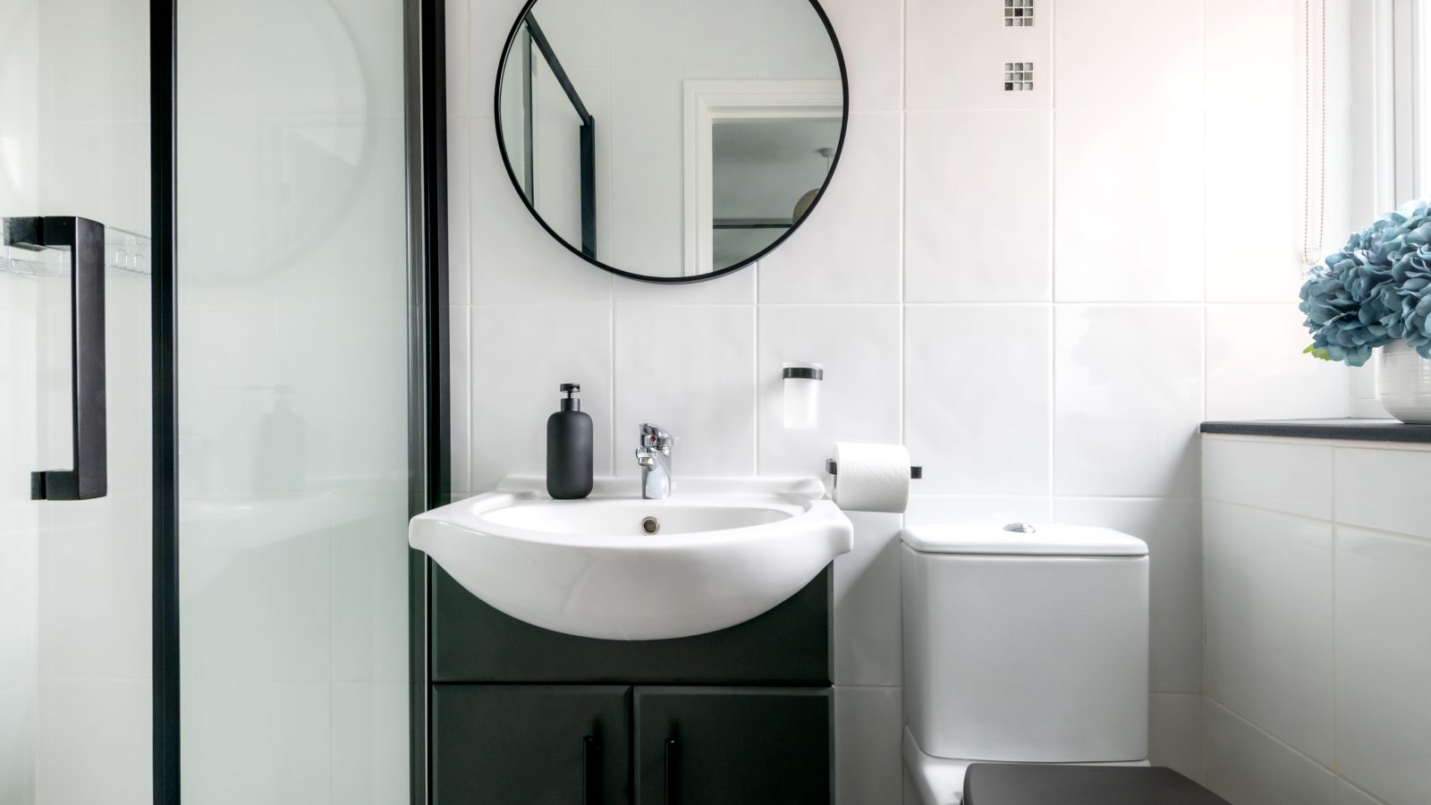 A modern West Acre bathroom with white tiled walls features a round black-framed mirror above a white sink. The sink cabinet is black, and a toilet is positioned next to it. A glass shower stall is visible on the left, and on the right, there's a window with a blue flower arrangement.