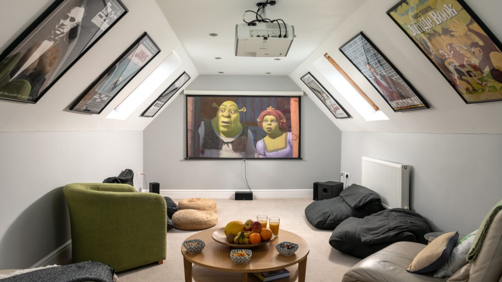 A cozy home theater in West Acre boasts a large screen showcasing an animated movie. The room is adorned with bean bags, a green armchair, and a leather couch. Movie posters grace the slanted walls, while a small round coffee table hosts snacks and drinks.