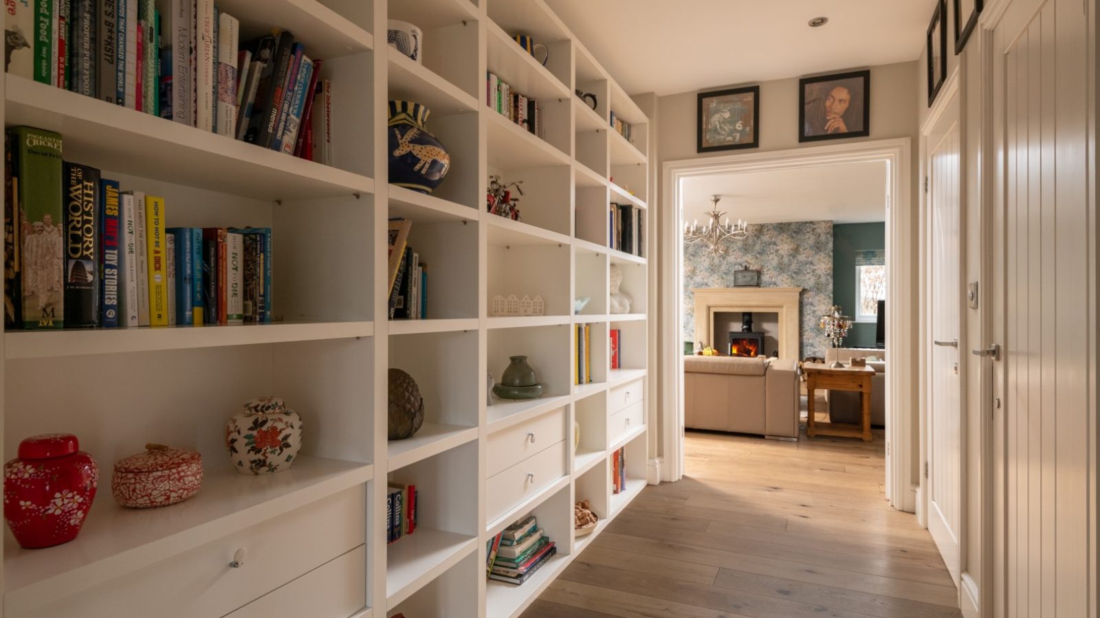 A hallway in West Acre features built-in white bookshelves on the left filled with books and decorative items. On the right are closed doors. The hallway leads to a living room with a fireplace, floral wallpaper, and a wooden floor. A ceiling light fixture illuminates the space.