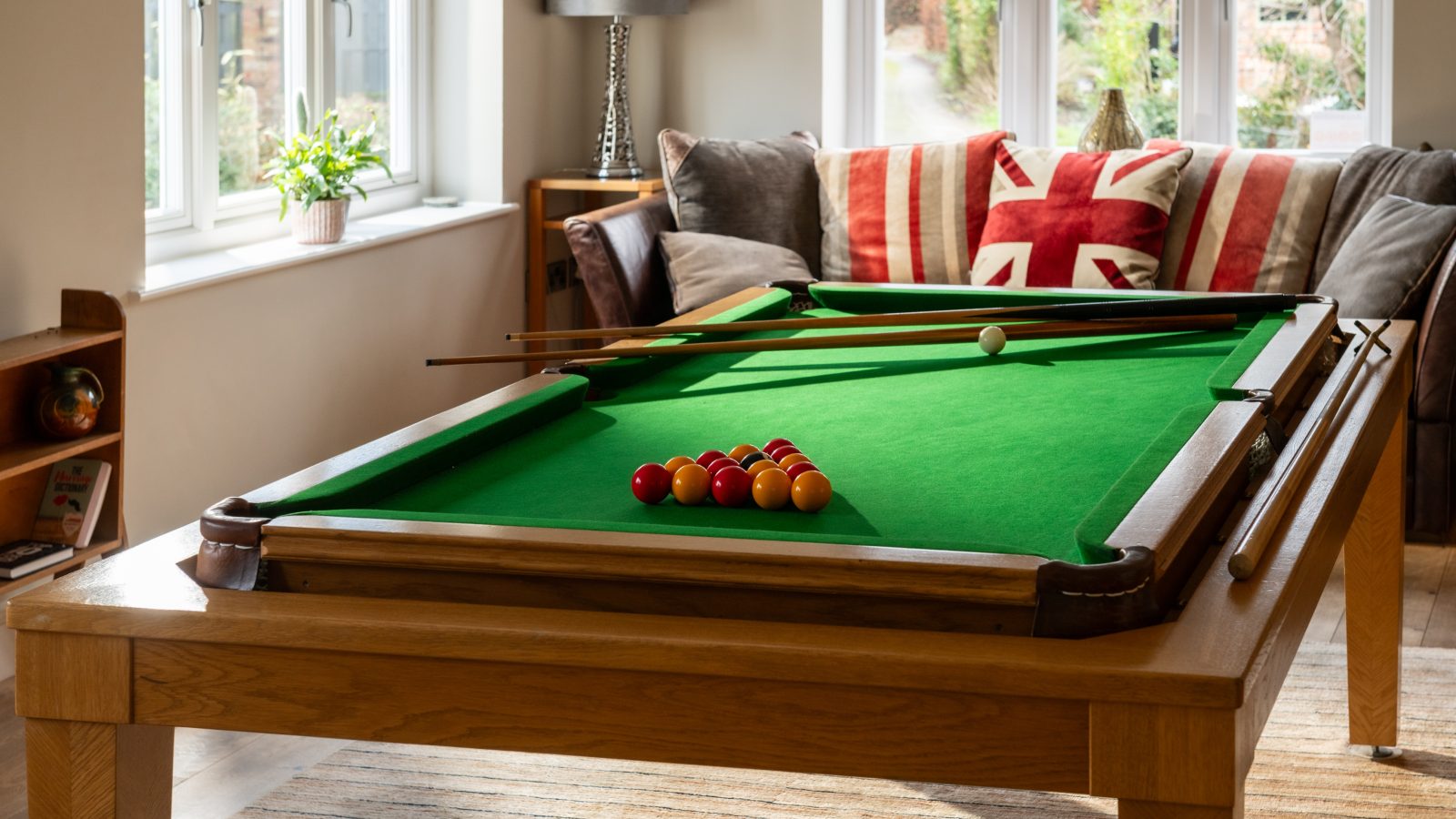 A cozy West Acre living room features a pool table centered with bright green felt, pool cues, and a rack of red and yellow balls ready for play. In the background, a gray couch adorned with Union Jack cushions sits near a large window that bathes the space in natural light.