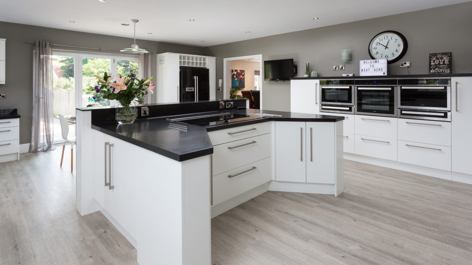 A spacious modern kitchen in West Acre features white cabinets and black countertops. An island with drawers and a vase of flowers is centered in the room. Multiple ovens, a large clock, and sleek appliances adorn the back wall. Light wood flooring and large windows enhance the brightness.