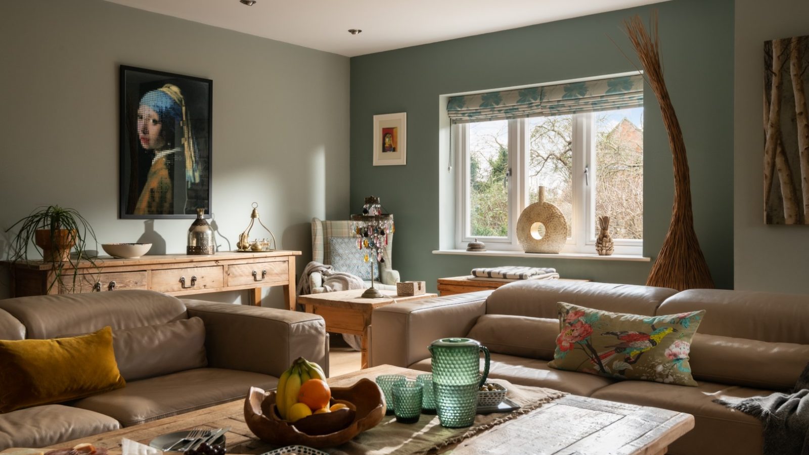 A cozy living room in West Acre with soft beige sofas adorned with colorful cushions. A rustic wooden coffee table holds a fruit bowl and other decor items. A large window lets in natural light, offering a serene outdoor view. Artwork and decorative items add character to the space.