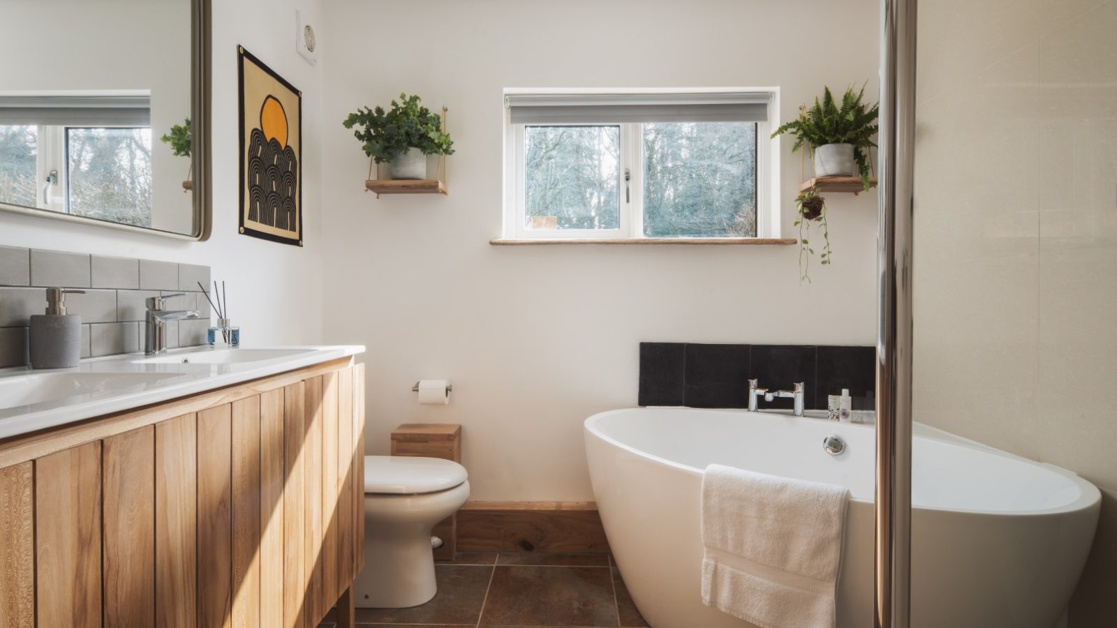 A modern, minimalist bathroom at Owl Lodge features a white freestanding bathtub, a wooden vanity with a white sink and large mirror, and a wall-mounted shower. Green plants on shelves and framed artwork adorn the walls while natural light streams through the window.