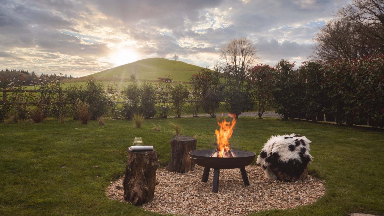 A cozy outdoor firepit surrounded by three tree stump seats—one with a fur cushion—set in the grass lawn of Owl Lodge. The peaceful background features a rolling green hill under a partly cloudy sky with the sun shining through, and a hedge borders the serene scene.