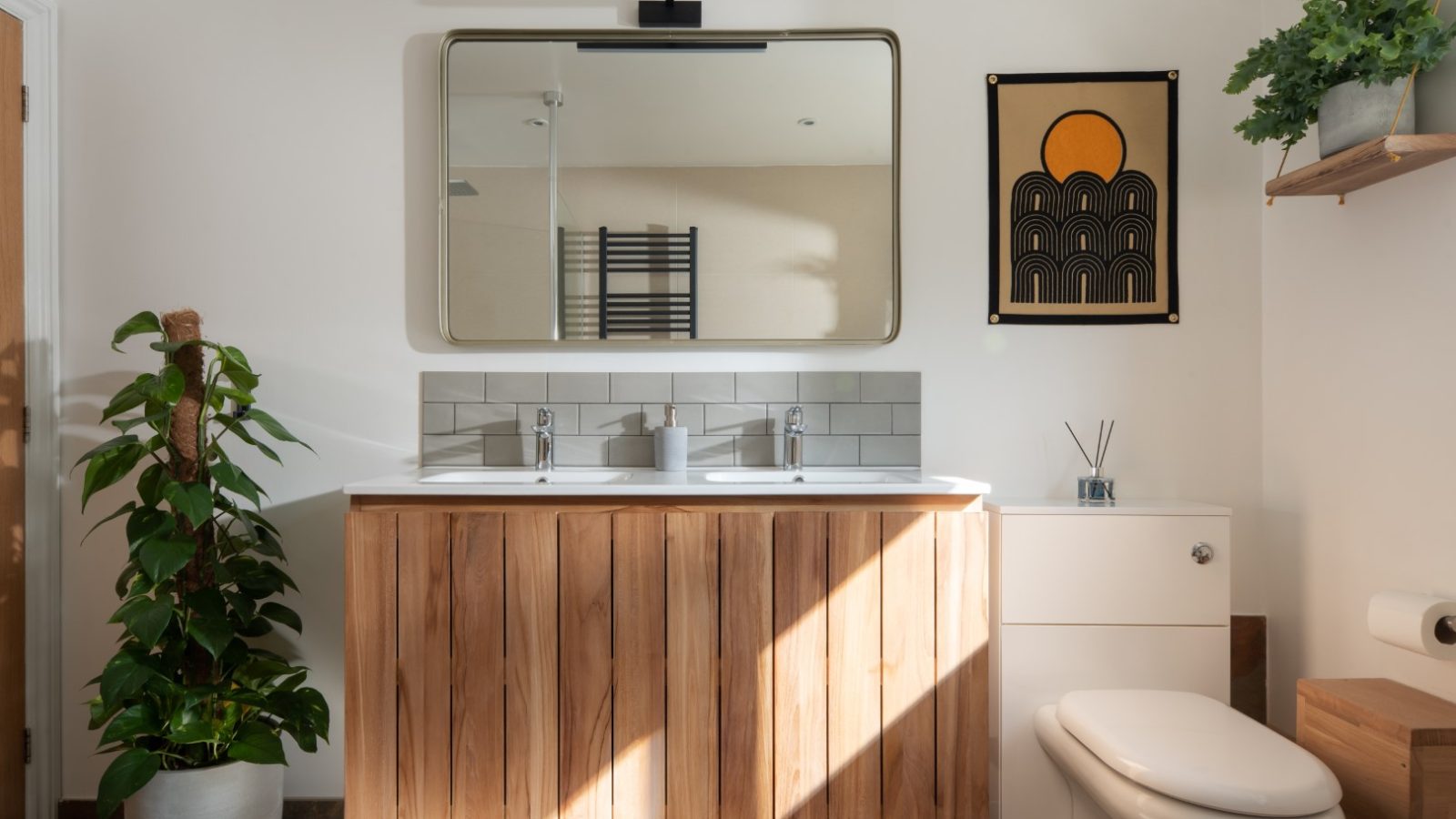 A modern bathroom at Owl Lodge features a wooden vanity unit with two sinks and a large mirror above it. A potted plant sits to the left, while a toilet is on the right. Above the toilet, abstract wall art depicting sun and arch designs adds a touch of flair.