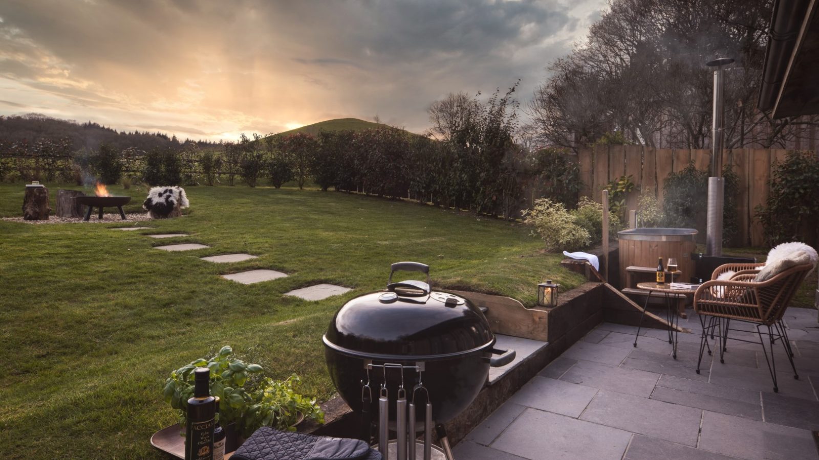 A backyard scene at sunset featuring a grill, a hot tub emitting steam, lounge chairs, a neatly manicured lawn, and a pathway of stepping stones. A fire pit with a bench and throw blanket sits nearby. Trees and bushes surround the area with rolling hills in the background, perfect for an Owl Lodge vacation stay.