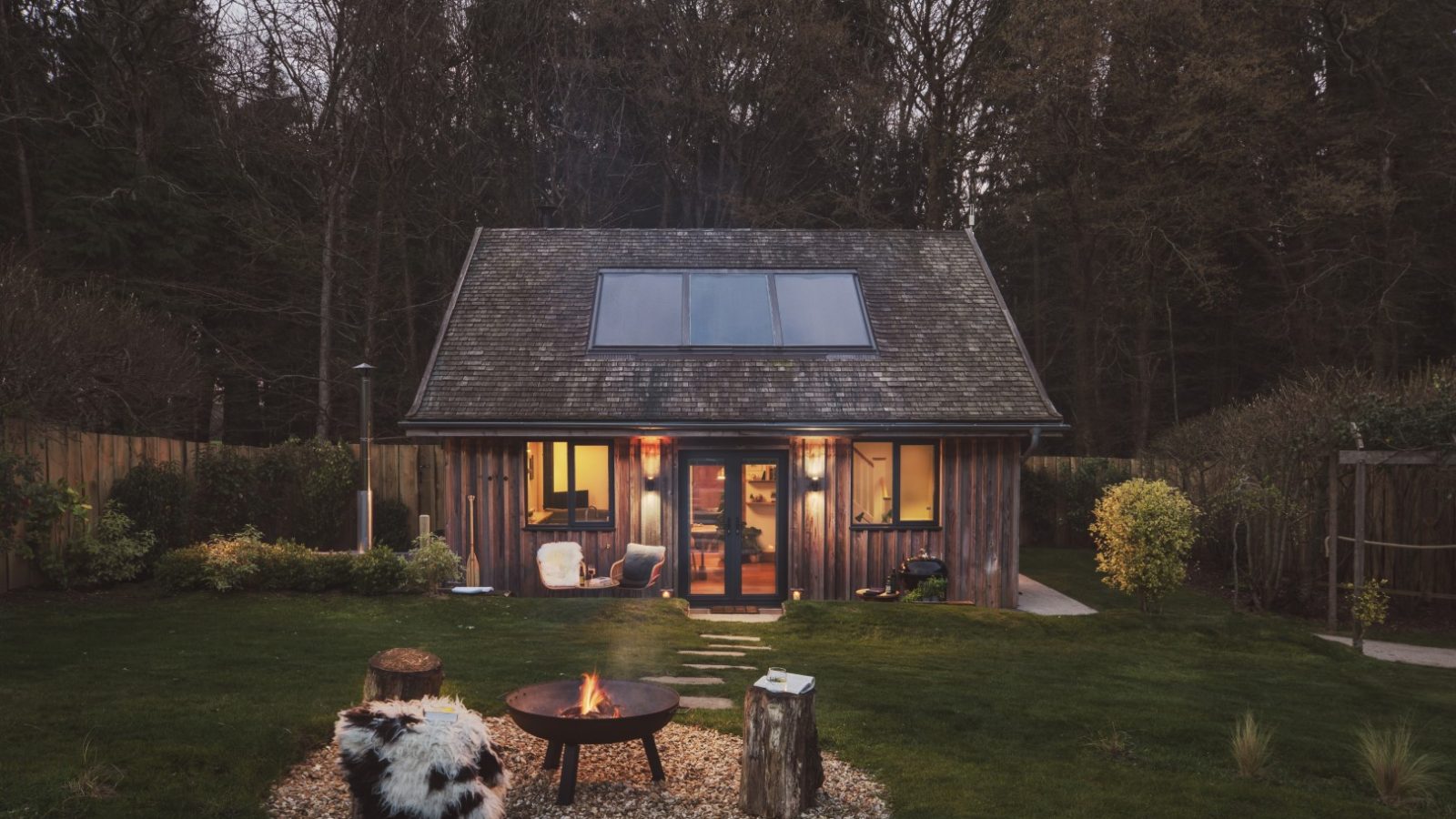 A cozy cabin with wooden siding and large windows is set in a grassy yard surrounded by trees. In front of the lodge, there's a fire pit with logs for seating. The yard includes a variety of plants, and an owl hoots softly as the evening sky suggests it's twilight.