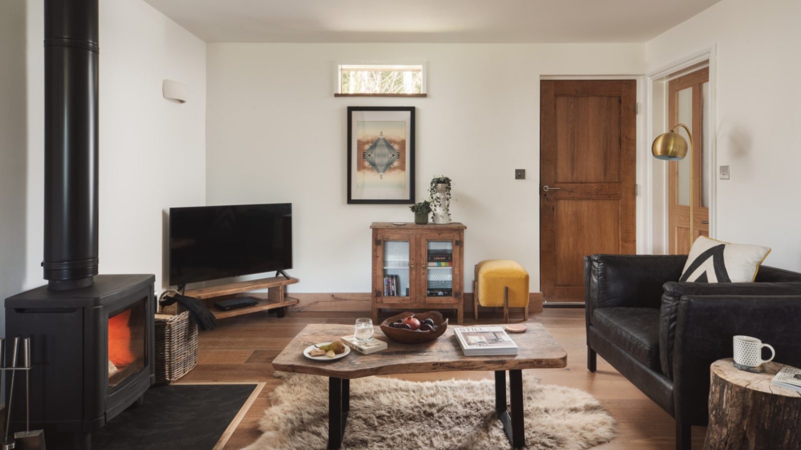 A cozy living room in Owl Lodge features a wood-burning stove on the left, a TV on a stand, and a wooden cabinet adorned with a plant and framed art above. A rustic coffee table with snacks and drinks sits on a fur rug between a black sofa and a yellow pouf. A door and lamp are in the background.