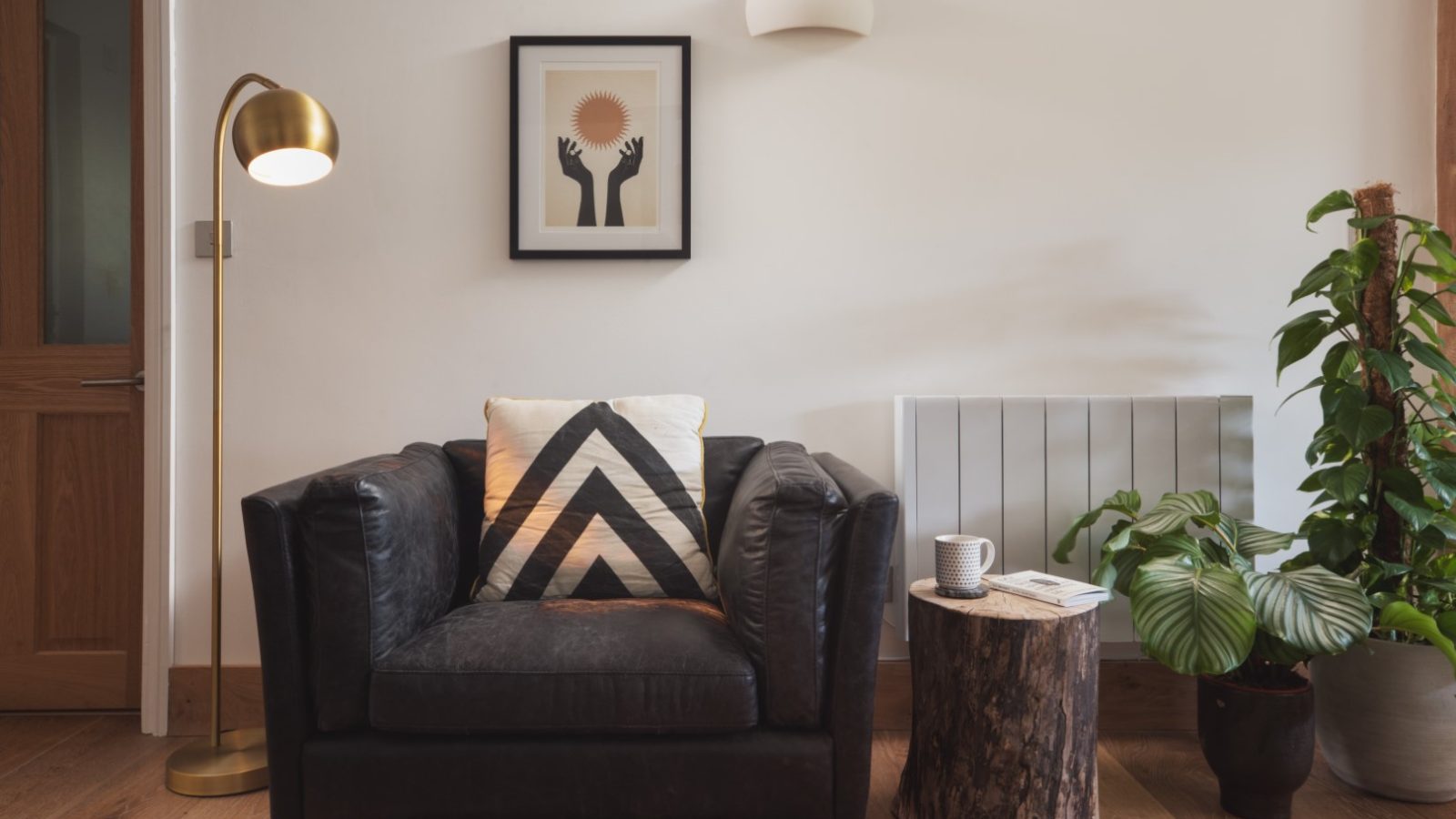 A cozy living room corner in the Owl Lodge features a black armchair with a geometric-patterned pillow, a wooden stump side table with a cup and book, a tall gold floor lamp, potted plants, and wall art depicting upright hands and a sun. The setting exudes warmth and comfort.