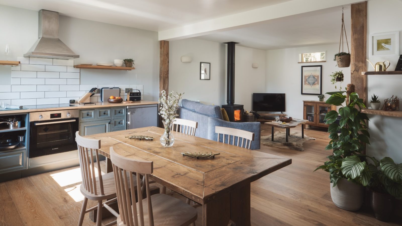 A cozy open-plan kitchen and living area with wooden floors evokes a charming lodge feel. The kitchen features modern appliances, light blue cabinets, and a subway tile backsplash. The dining area has a rustic wooden table with four chairs, while the living area includes a sitting area adorned with plants, an owl ornament, and a TV.