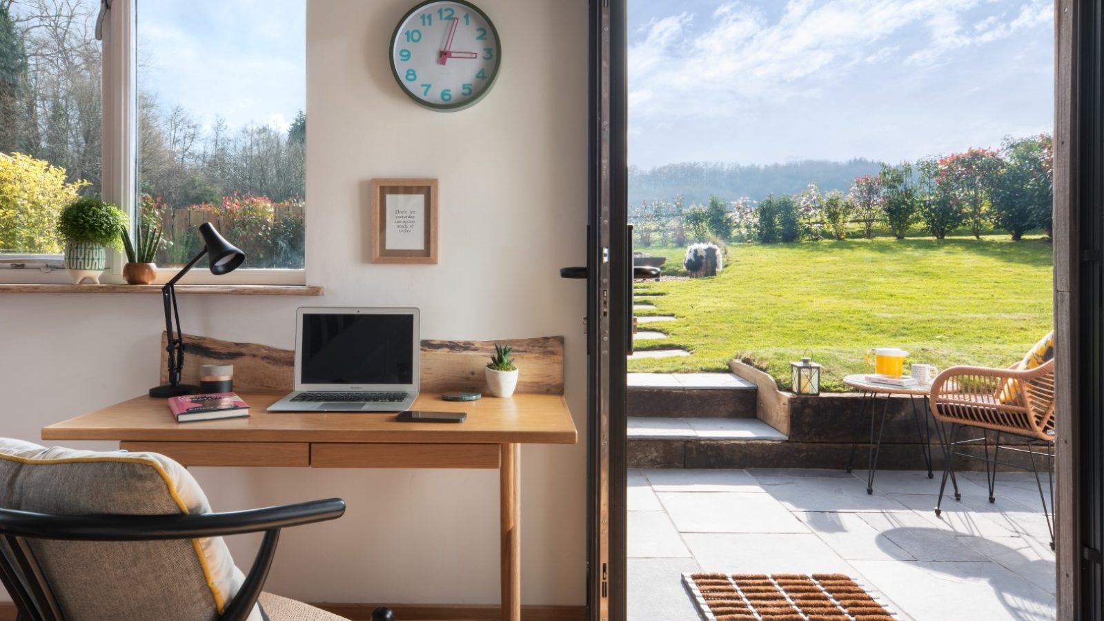 A serene home office at Owl Lodge features a window and open door showcasing a lush green backyard. The desk holds a laptop, lamp, and books. A chair with a cushion faces the desk, and a clock and framed quote hang on the wall. Sunlight floods the room, creating a calming ambiance.