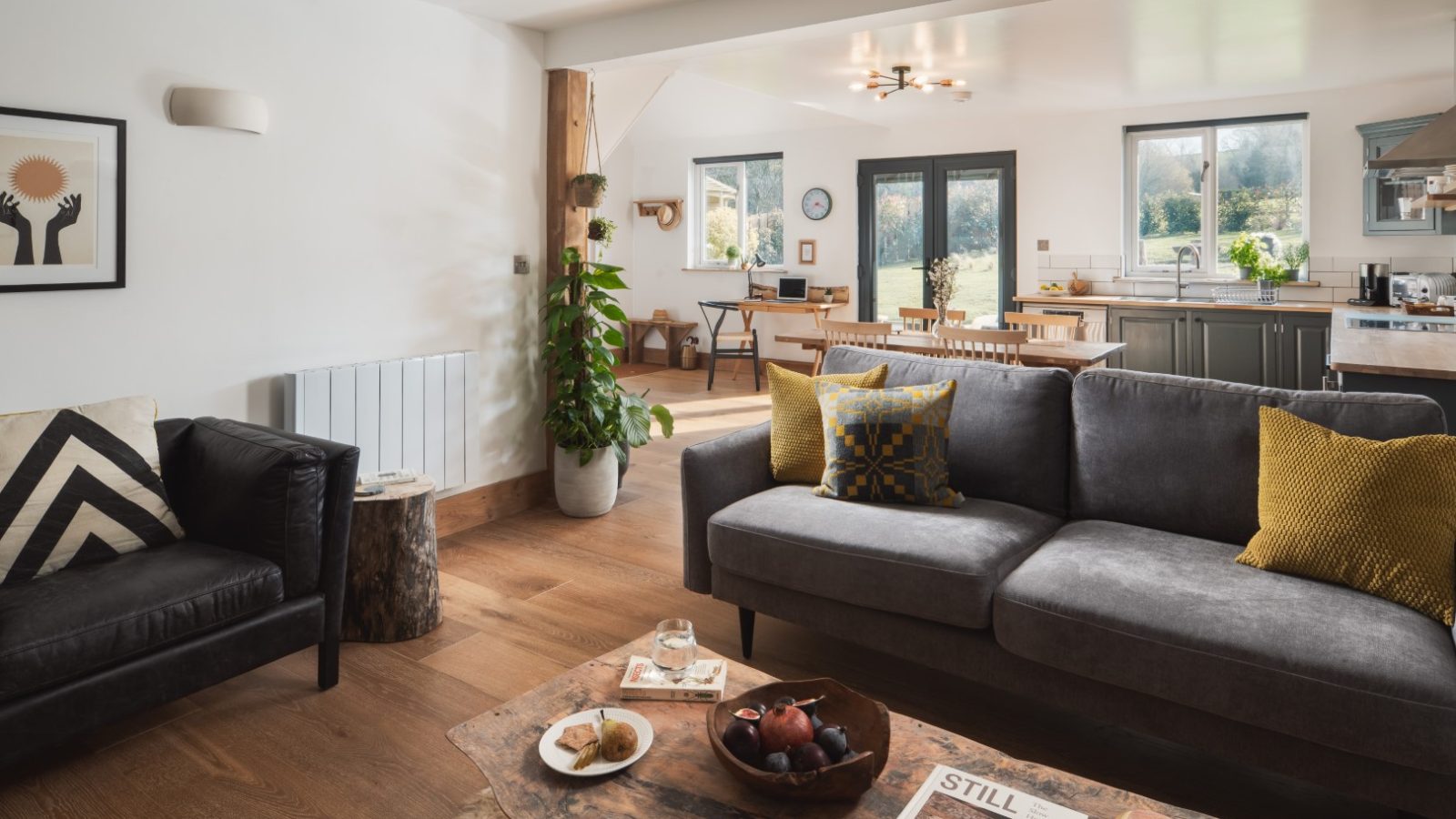 A living room with a black leather couch, gray fabric sofa with yellow pillows, a wooden coffee table, and a potted plant. The area transitions into the Owl Lodge's kitchen and dining space with wooden chairs, large windows, and modern decor. Warm, natural lighting fills the room.