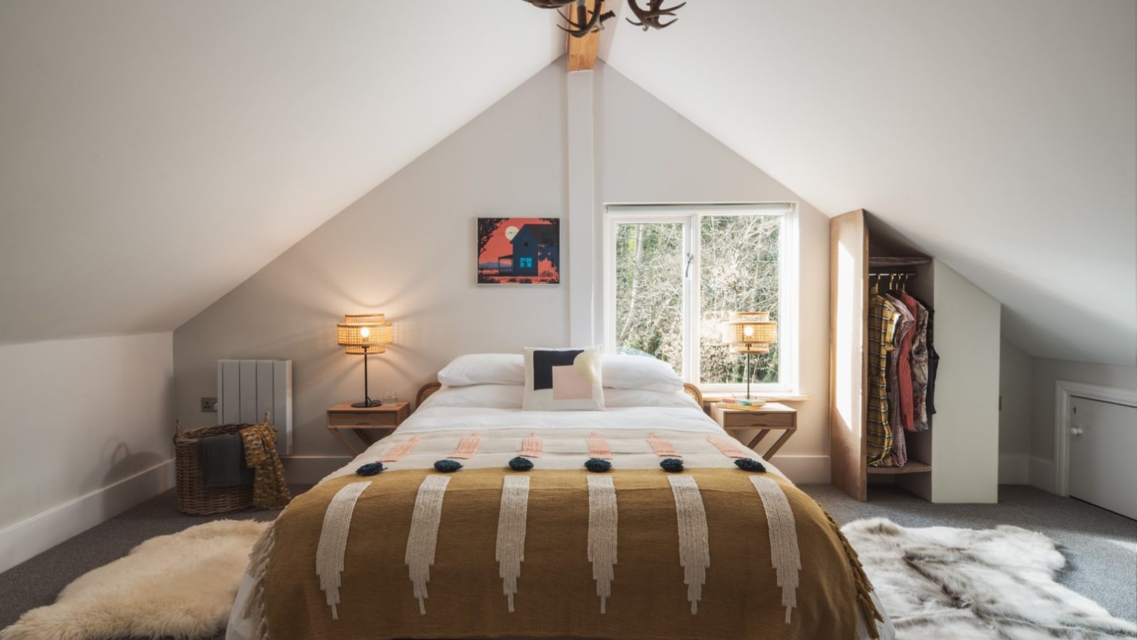 A cozy attic bedroom with a sloped ceiling features a bed adorned with patterned bedding. Flanked by wooden nightstands with lamps, the bed sits below a window that brings in natural light. To the right, an open closet displays hanging clothes, adding to the rustic charm of this Owl Lodge retreat.