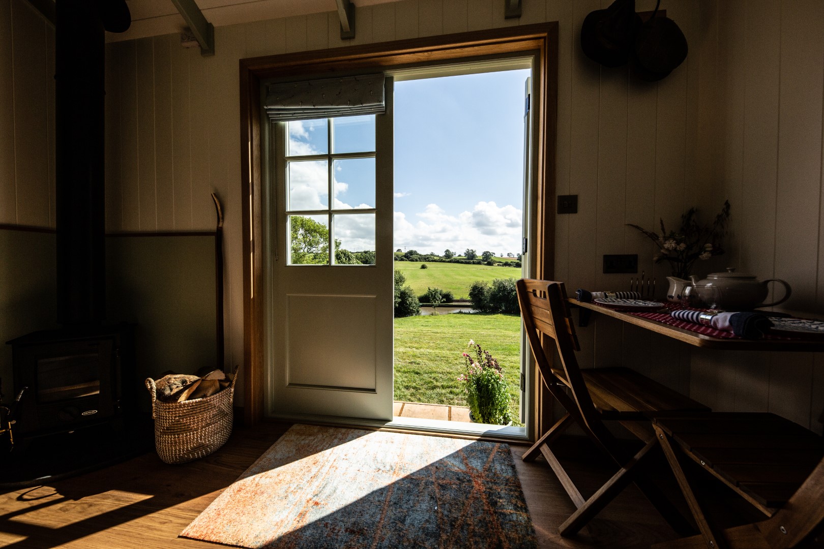 A cosy room with light streaming through an open door that leads to a picturesque countryside view, just 2 hours from London. The room features a wood-burning stove with firewood in a basket beside it, a rustic dining table set, and a bright rug partially on the wooden floor—one of the ideal places to stay near London.