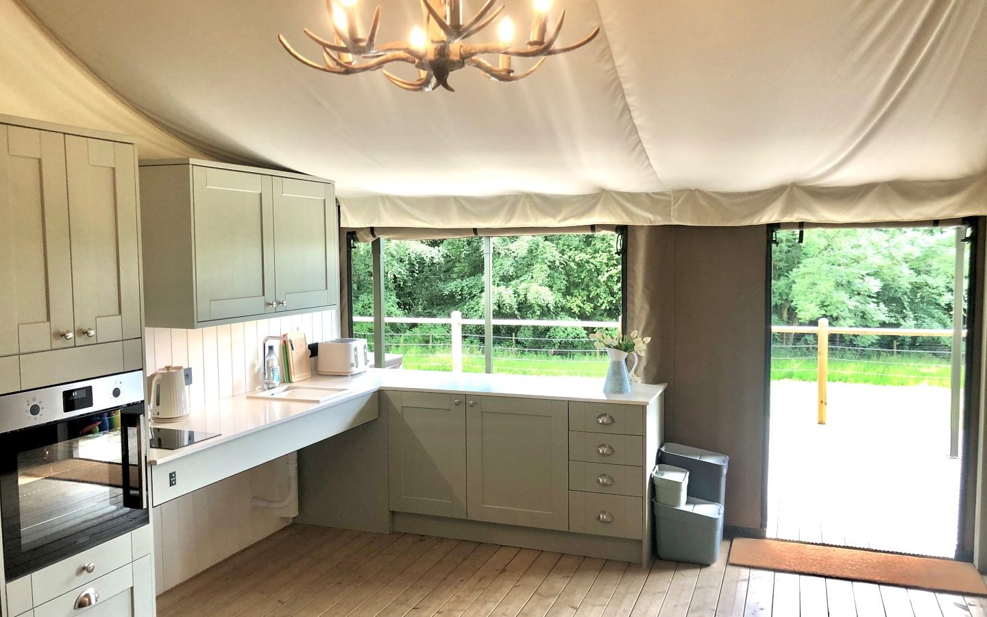 A spacious, bright kitchen with light green cabinets and wooden flooring. It features modern appliances, including an oven, kettle, and countertop. A chandelier with an antler-style design adds a rustic touch. Large windows and a glass door provide a view of the lush flora outside.