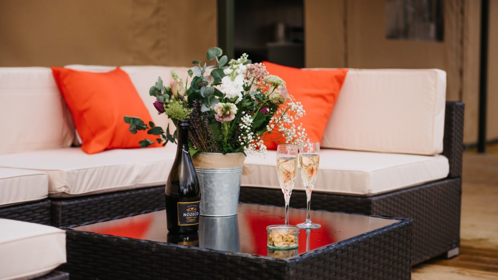 A chic patio setup with a wicker coffee table adorned with a floral centerpiece, a bottle of sparkling wine, two filled wine glasses, and a small bowl of snacks. The background features a white sectional sofa with vibrant orange cushions and vibrant flora enhancing the ambiance.