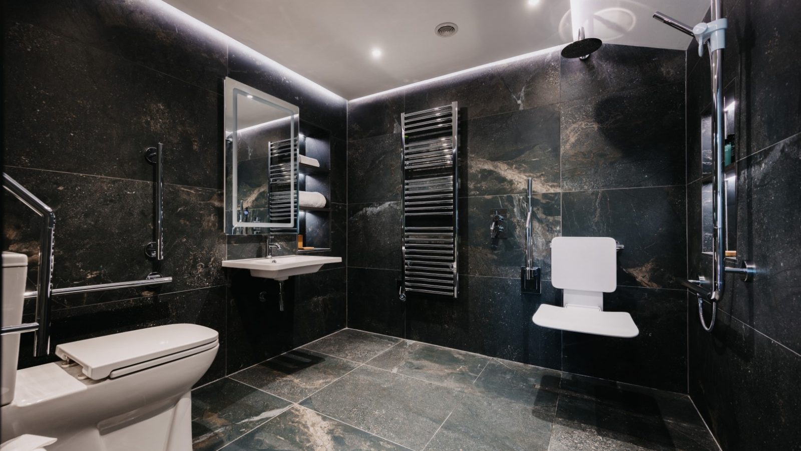 A modern bathroom with dark stone tile walls and floors, featuring a toilet, a wall-mounted sink with a rectangular mirror, a heated towel rack, a wall-mounted shower chair, and a rainfall showerhead. The space is accented with flora to bring in natural freshness. The lighting is recessed in the ceiling.