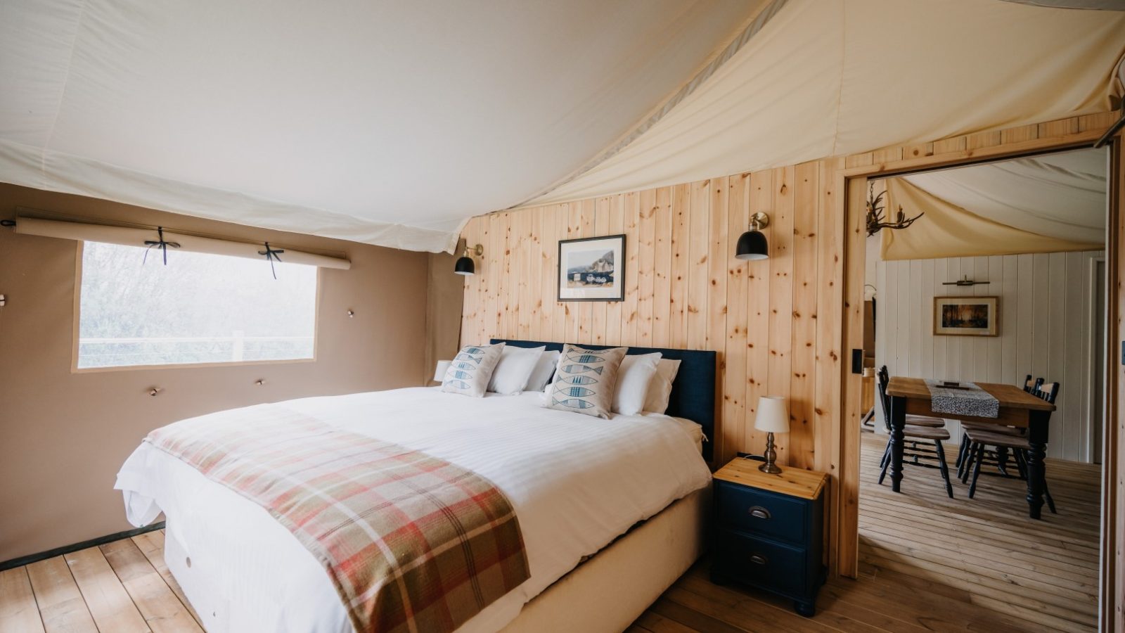 A cozy cabin bedroom with a large bed covered in a white duvet and plaid blanket. The room features light wood paneling, two black wall-mounted lamps, and a framed picture of local fauna above the bed. In the background, a dining area with a wooden table and chairs is visible.