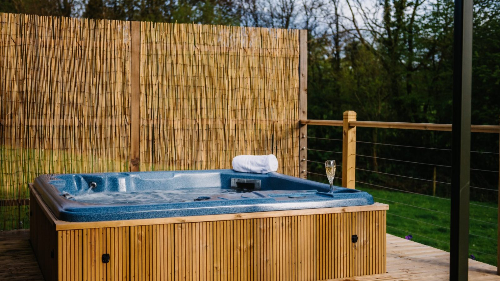 A wooden deck with a blue outdoor hot tub filled with water, situated next to a wooden privacy screen. A rolled-up white towel and a glass of champagne are placed on the edge of the hot tub. Lush green flora frames the background, enhancing the tranquil setting.