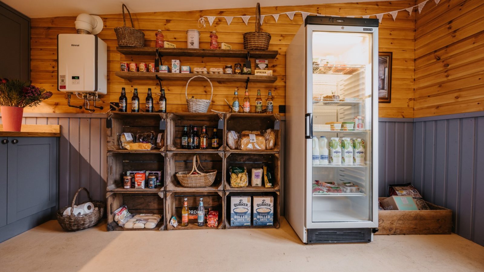 A cozy pantry with wooden shelves stocked with various food items, including canned goods, snacks, and drinks. A glass-display refrigerator contains milk and other refrigerated products. Decorative baskets, bunting, and flora motifs add a homey touch to the wooden walls.