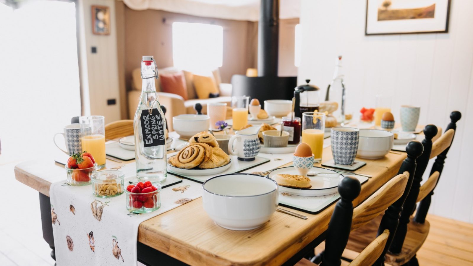 A wooden dining table set for breakfast with plates, cups, glasses of orange juice, a bottle of water, pastries, boiled eggs, berries, and condiments. The animal print table runner adds a touch of flora and fauna to the cozy, well-lit room with a sofa and framed picture in the background.