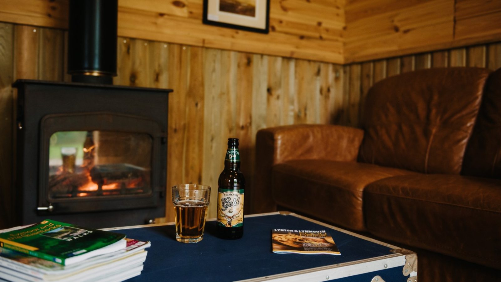 A cozy cabin interior with a lit fireplace. A glass and bottle of beer sit on a blue wooden trunk used as a coffee table, surrounded by magazines. The brown leather couch is partially visible to the right, while the wooden walls and touches of flora add to the rustic, nature-inspired decor.