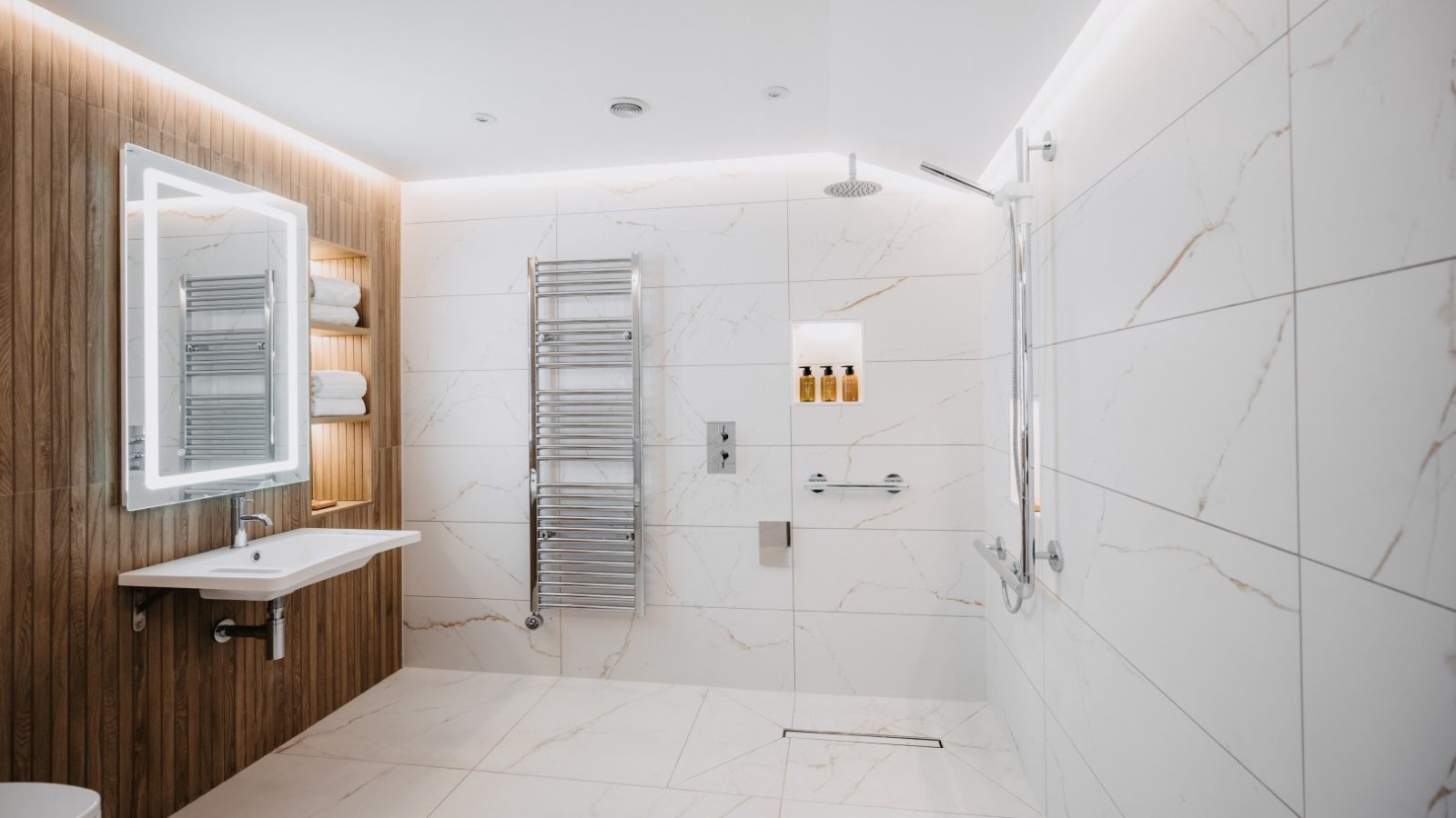 A modern bathroom with white marble tiles, a large illuminated mirror, a white sink, and a wooden accent wall. Features a towel rack, a walk-in shower with a rain showerhead, and wall-mounted toiletries. The space is well-lit with recessed ceiling lights and touches of fauna-inspired decor.