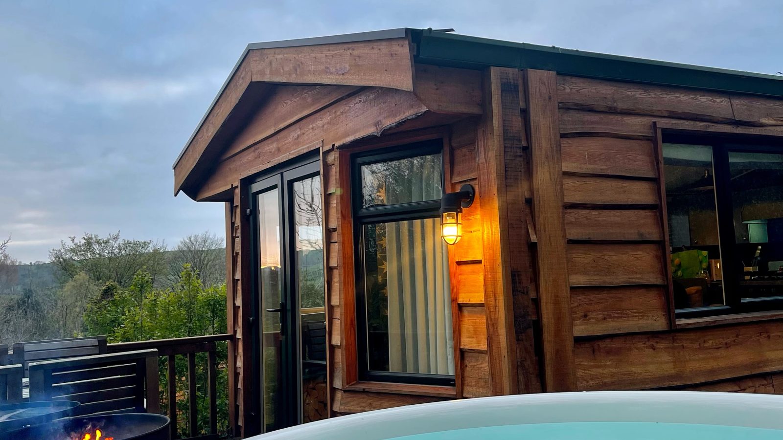 A serene scene of an outdoor wooden cabin at dusk at Howgill Lodge, with a hot tub in the foreground filled with clear blue water. Next to it is a small outdoor fire pit emitting smoke. The cabin features a large window and a lit wall lantern. The sky is overcast.